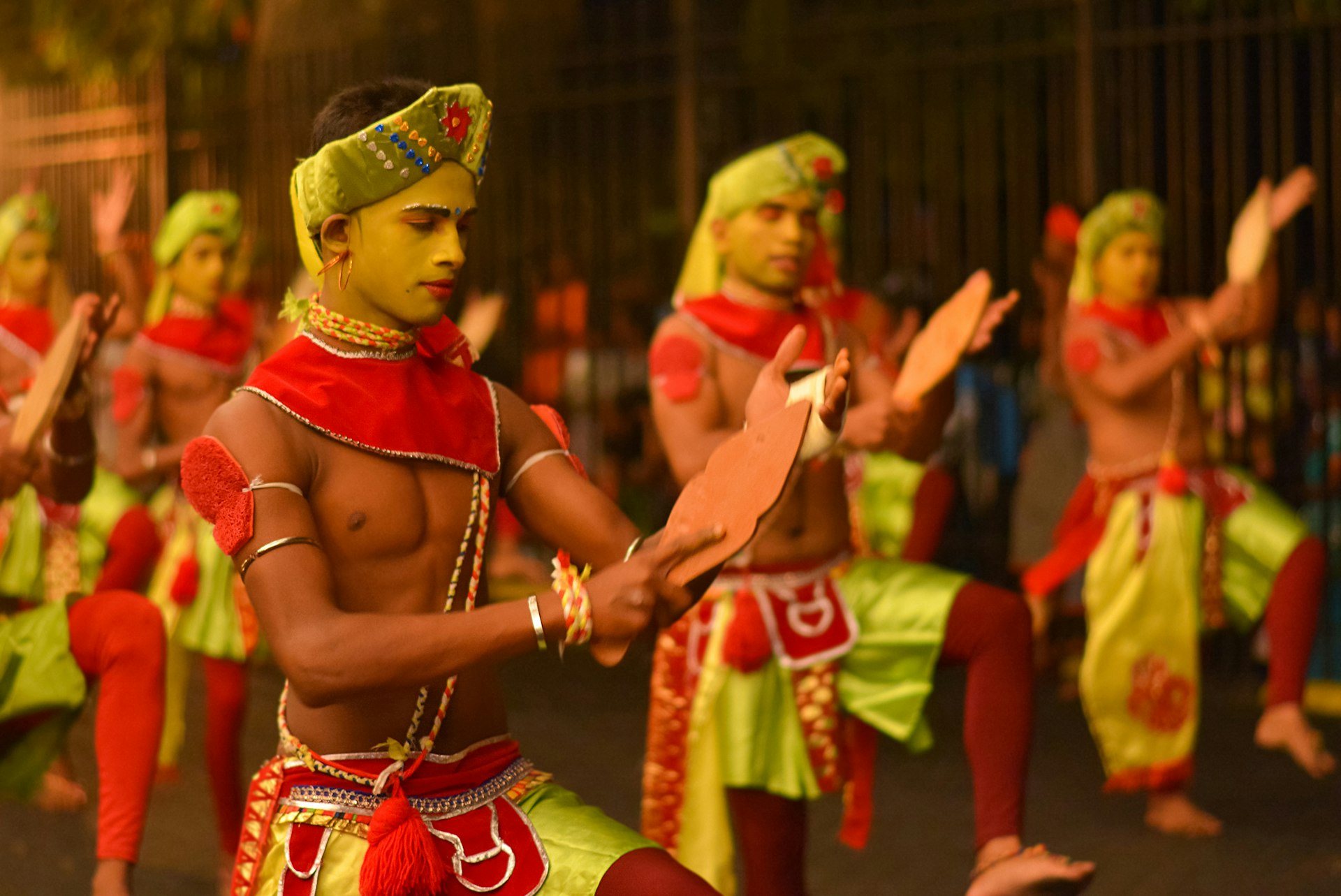 A troupe of male dancers in costume and make up perform together in a row, clapping their hands agains a wooden paddle