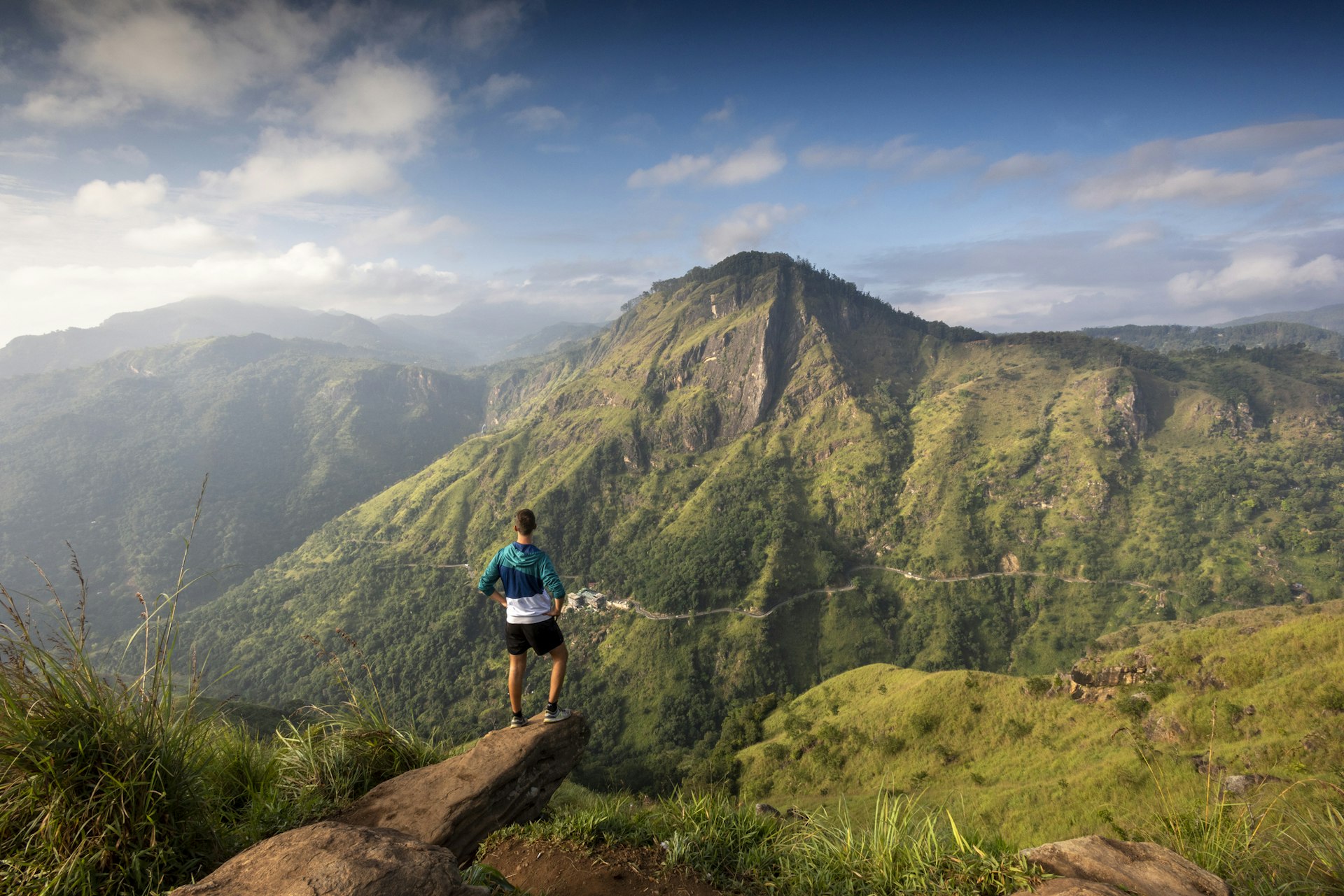 A man stands on a peak looking towards another distinctive hill in a lush green region