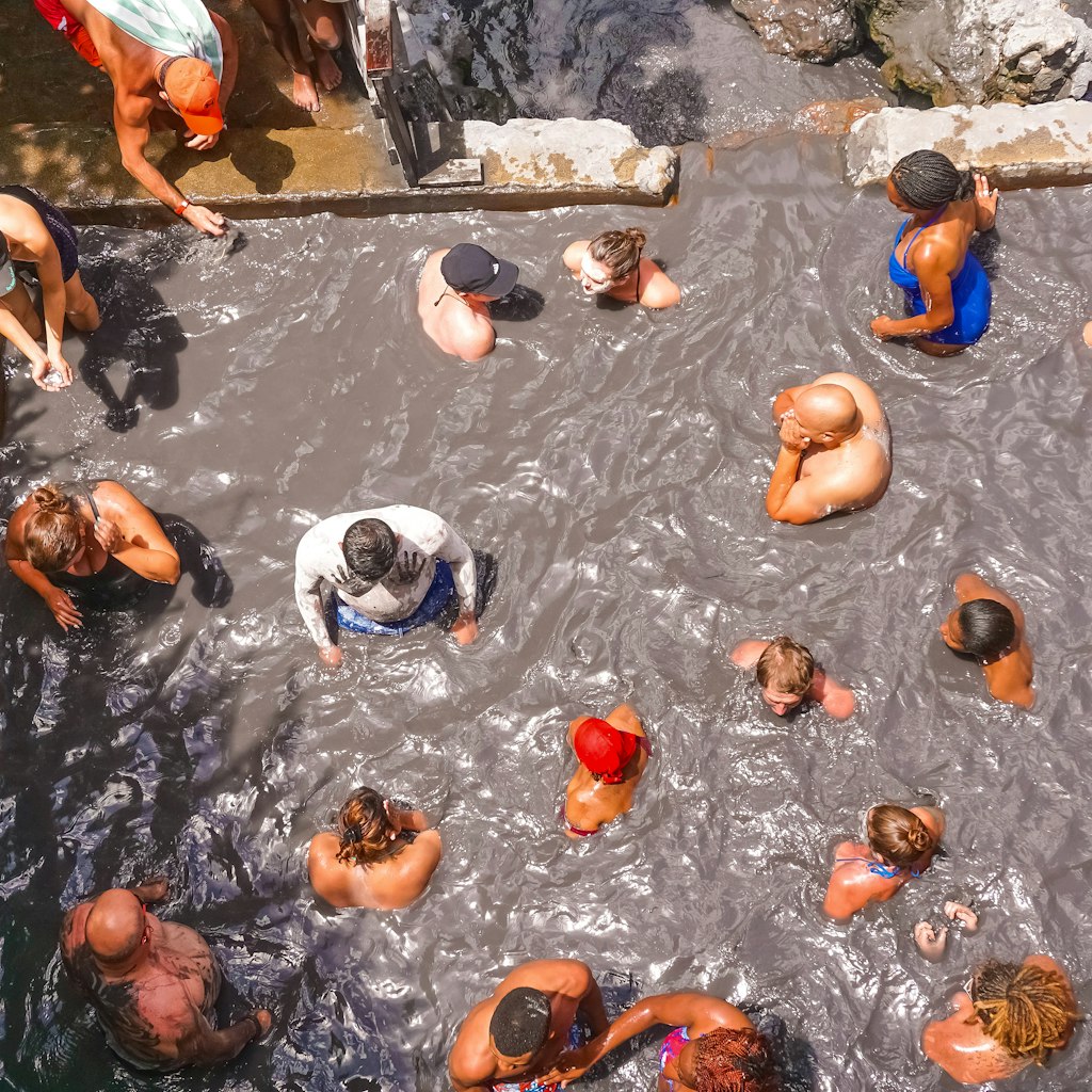 Aerial shot of a group of people sitting in a large stone pool filled with gray mud at Sulphur Springs in St Lucia. 