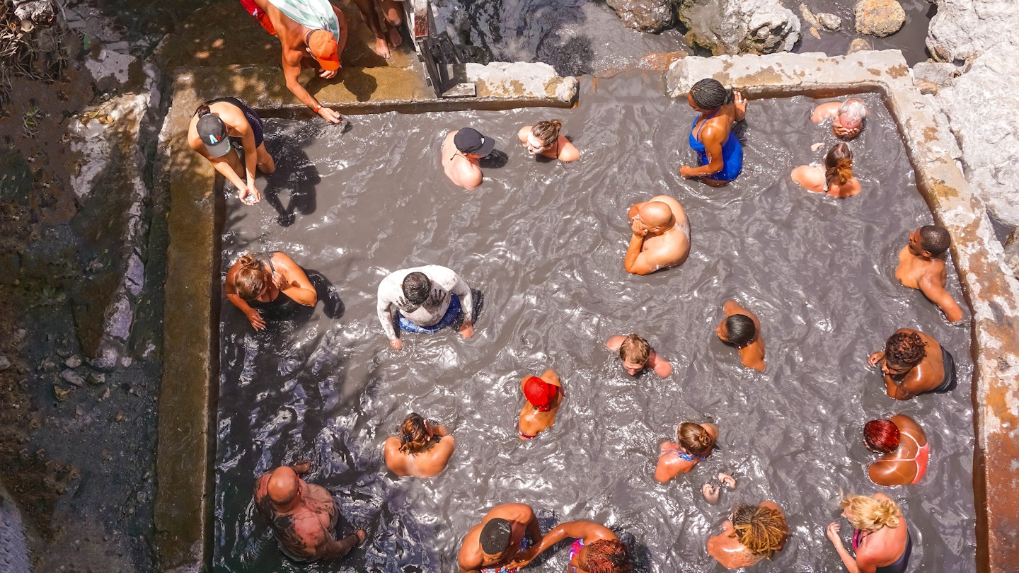 Aerial shot of a group of people sitting in a large stone pool filled with gray mud at Sulphur Springs in St Lucia. 