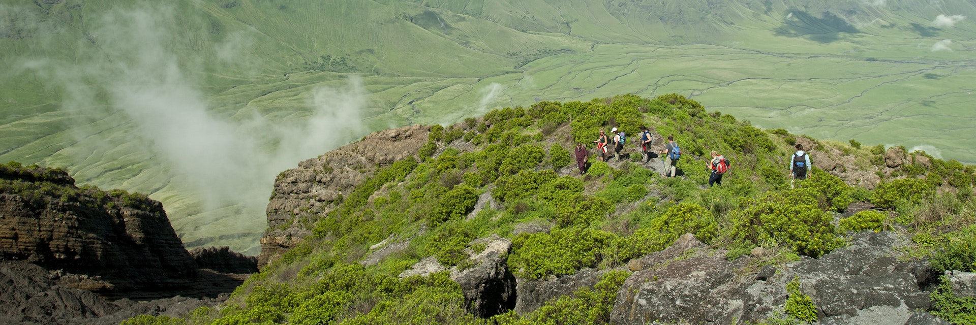 Ol Doinyo Lengai, Tanzania - January 1, 2007: A group of tourists is hiking down from the  top of Ol Doinyo Lengai into the green Rift Valley, in the background the Escarpment of the Rift Valley is visible. In the foreground there is some volcanic smoke coming out of a crack.