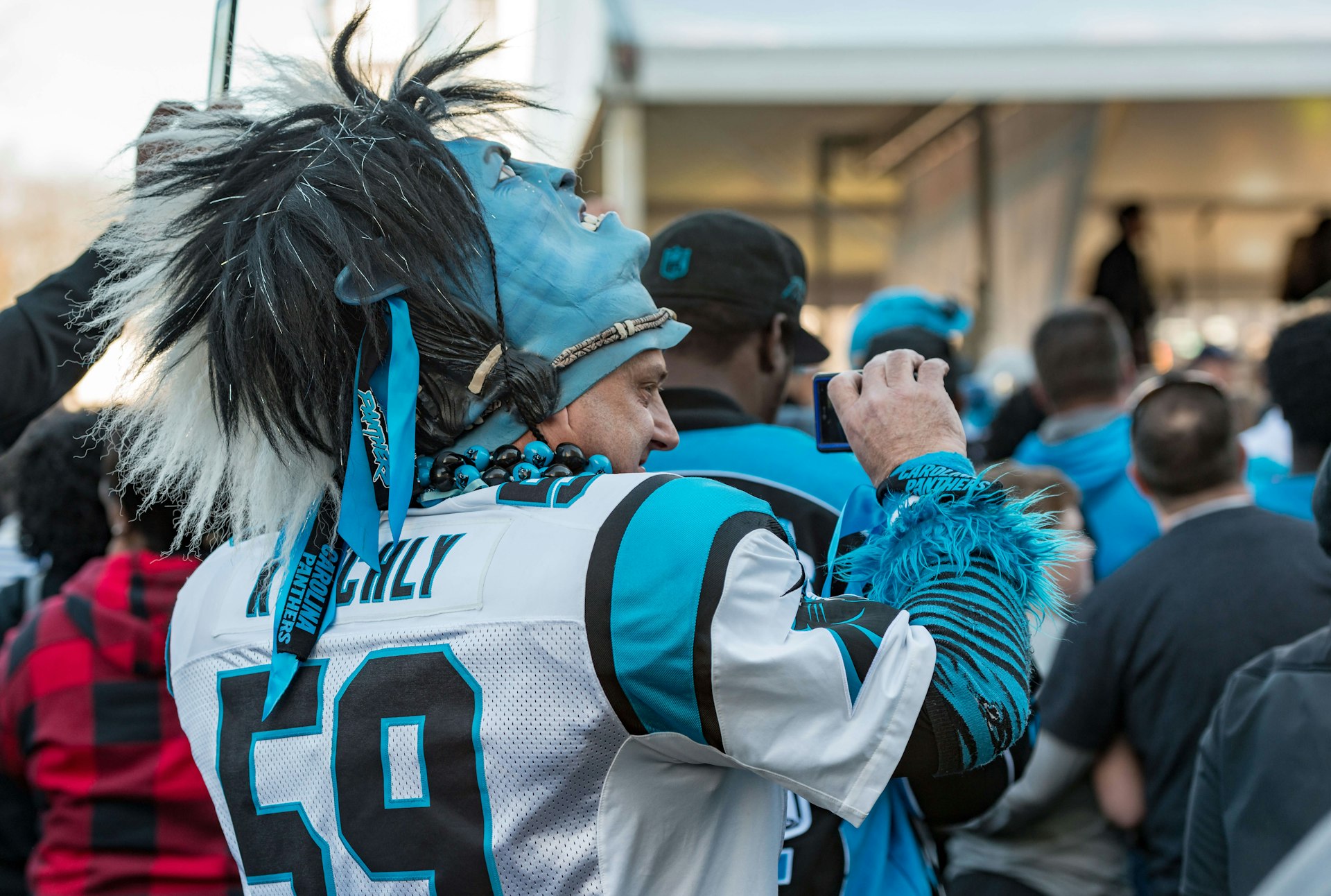 Closeup of a Carolina Panthers football fan, wearing a jersey and a mask pushed back on his head, taking a picture