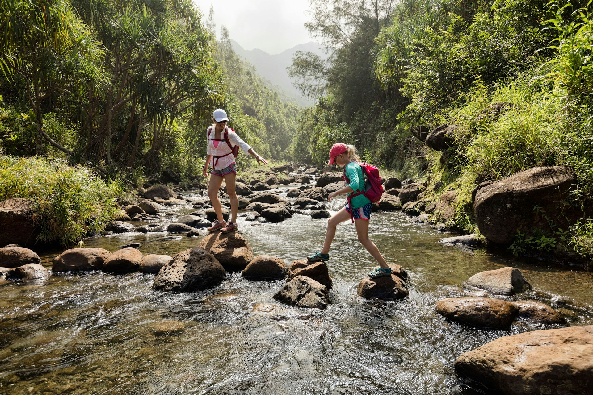 Mother and daughter crossing a creek via stones in a lush rainforest, Na Pali Coast, Kaua’i, Hawaii