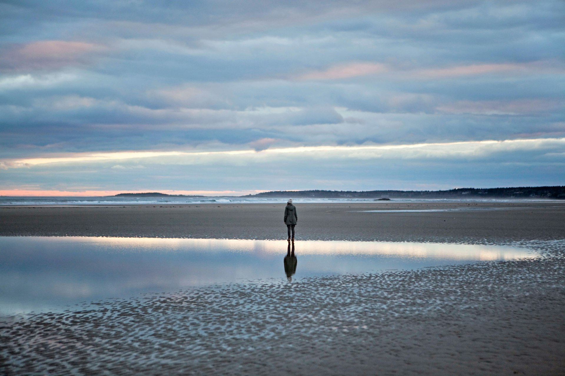 A woman stands reflected in a tidal pool on the beach in Maine