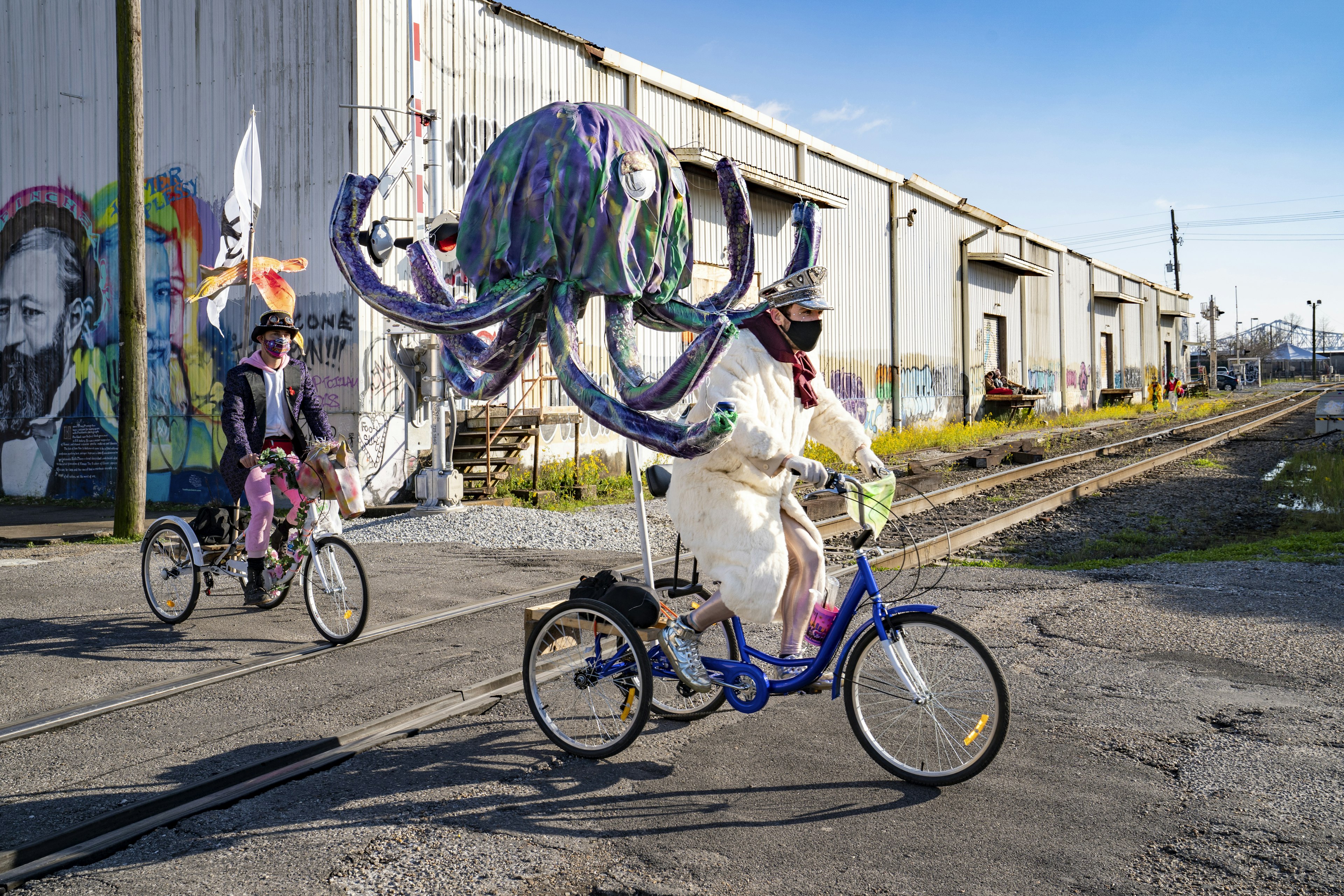 Costumed Mardi Gras revelers ride bikes through the Bywater in front of street art.