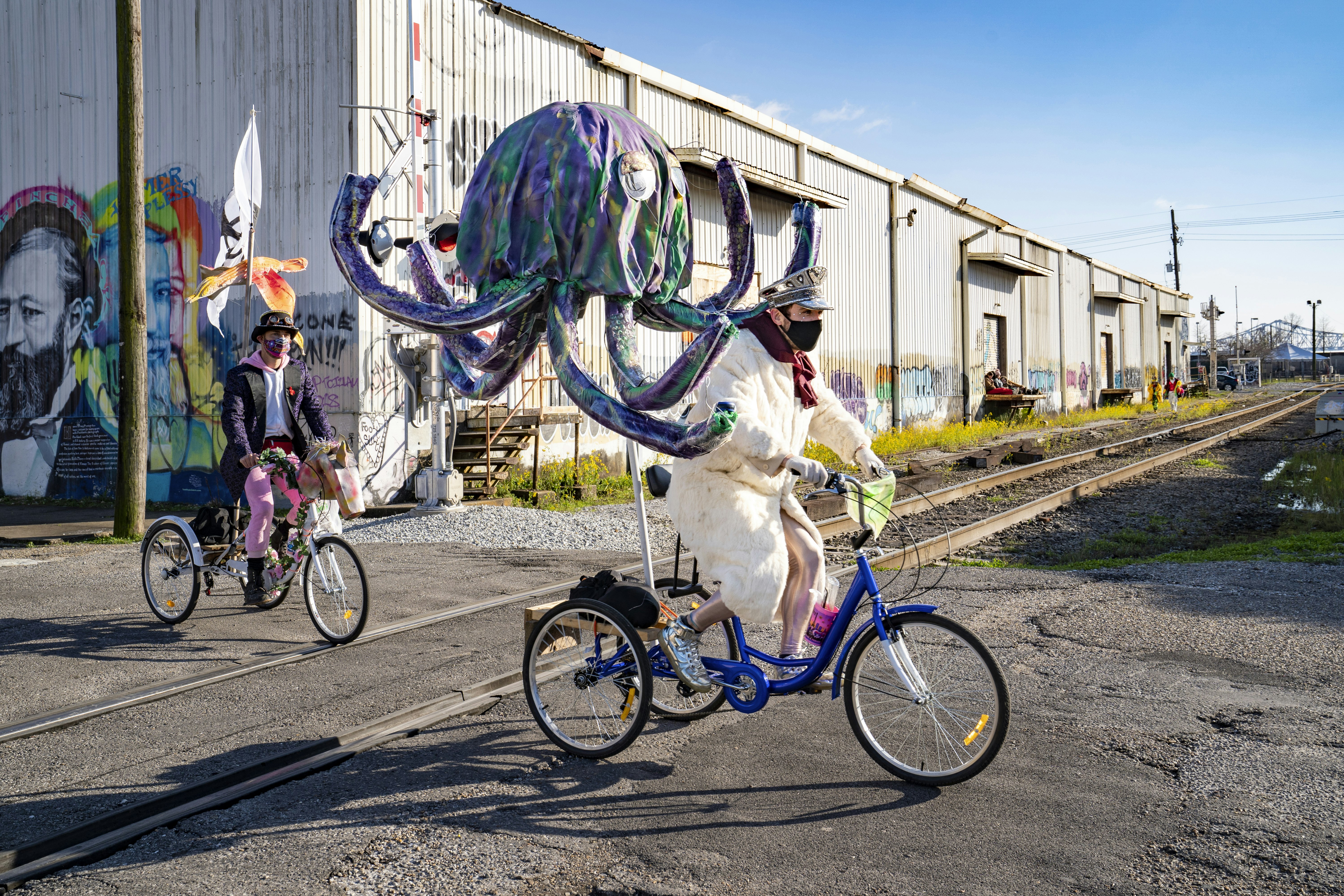 Costumed Mardi Gras revelers ride bikes through the Bywater in front of street art. 