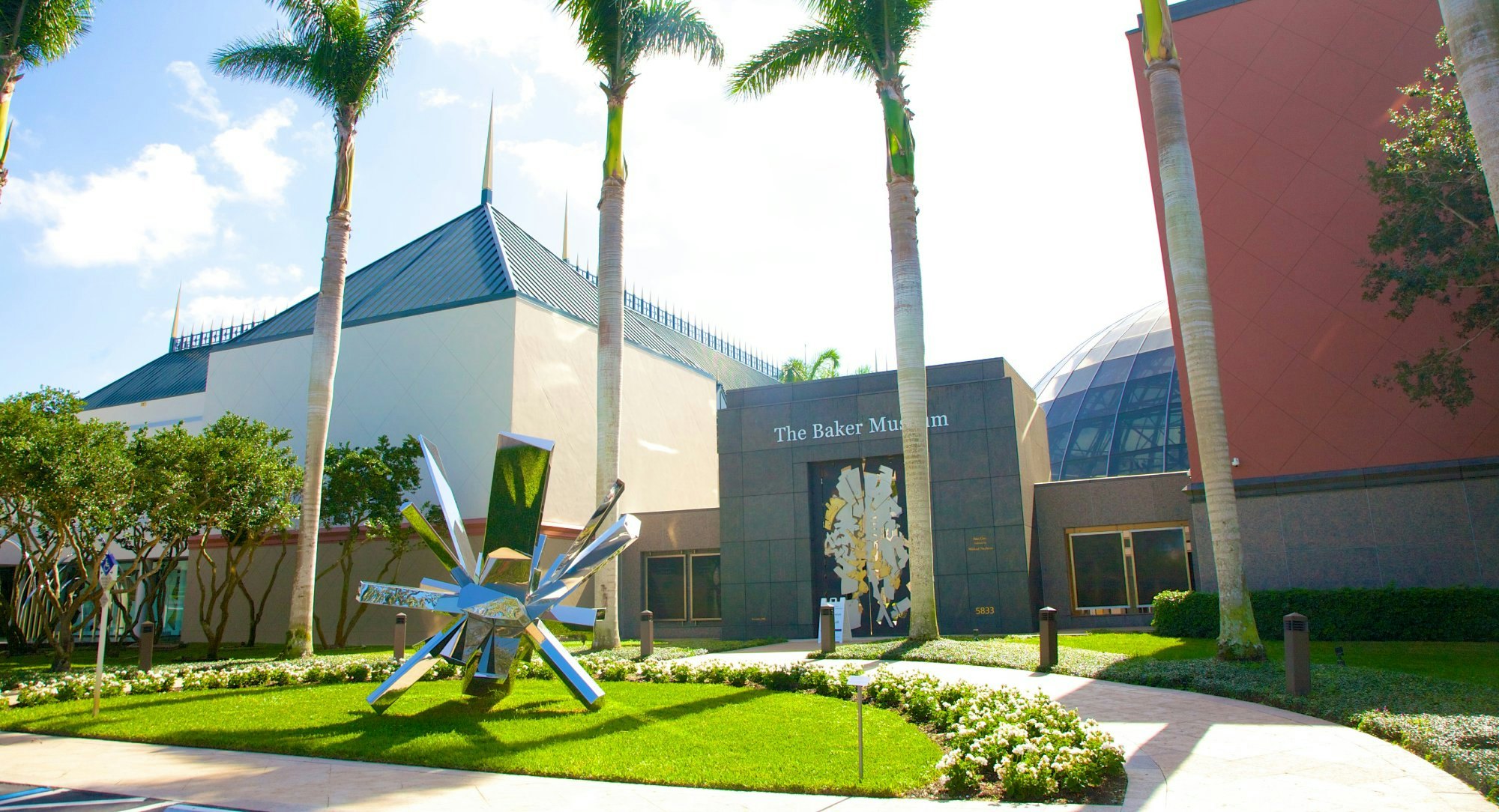 Stainless steel sculpture by artist Arik Levy titled RockGrowth 370, from 2017, in front of palm trees and main entrance to The Baker Museum, a modern art museum in Naples, Collier County, Florida, USA.  The museum houses a diverse collection of art in a three-story, 30,000-square-foot facility. Permanent collections include American modernism, 20th-century Mexican art, sculpture and 3-dimensional art.  The building, with its angled brick- and stone-toned fascia tiles, was designed by Florida architect Gene Aubry, who also designed the Hayes Hall building next to it.