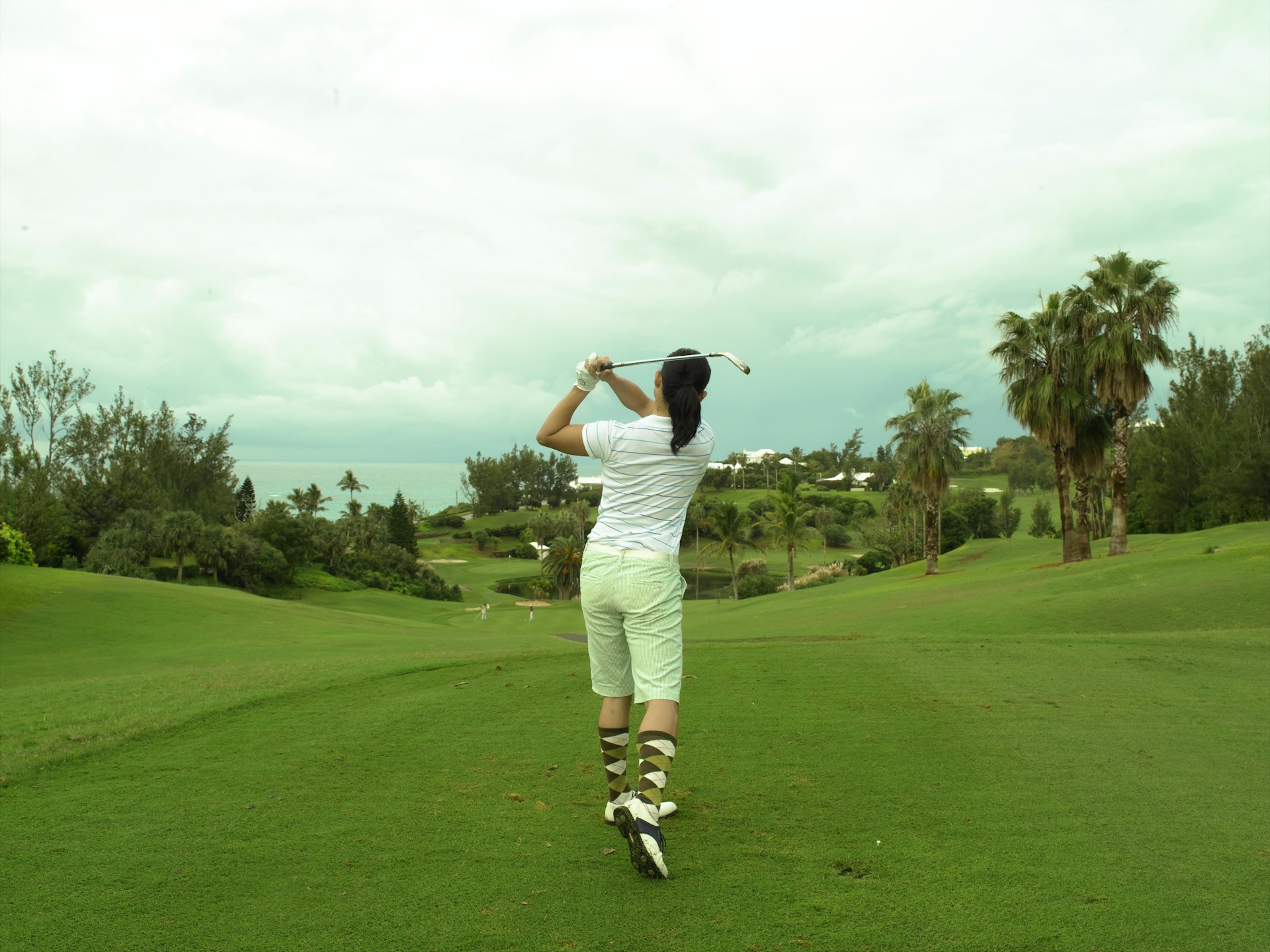 A woman takes a swing on a golf course in Bermuda. It's a cloudy day. 