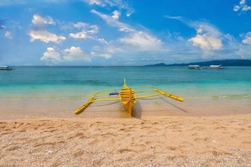 A traditional Filipino wooden outrigger boat in shallow turquoise water and picturesque sandy beach, in the tourist resort of Puerto Galera, Philippines.