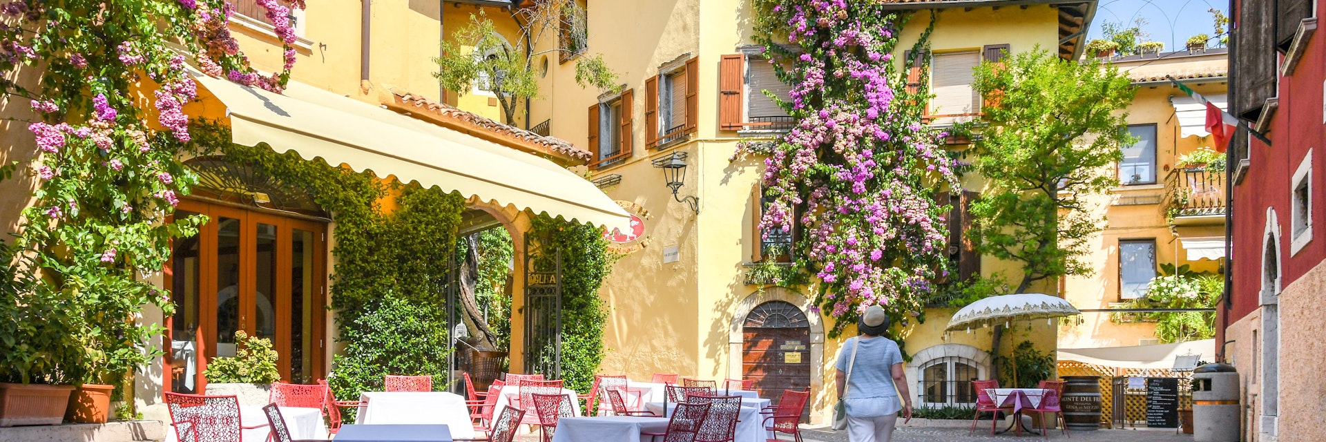 Gardone Riviera, Lake Garda, Italy - September 2018: Person walking past a restaurant with outside tables in Gardone Riviera. The buildings are covered by large flowering climbing plants.