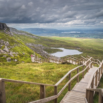People enjoying a walk on steep stairs of wooden boardwalk in Cuilcagh Mountain Park with a view on lake and valley below, Northern Ireland