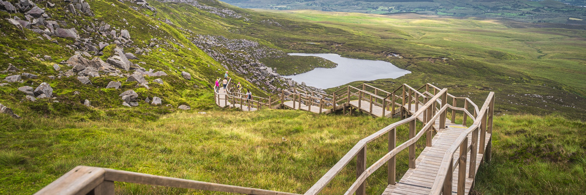 People enjoying a walk on steep stairs of wooden boardwalk in Cuilcagh Mountain Park with a view on lake and valley below, Northern Ireland