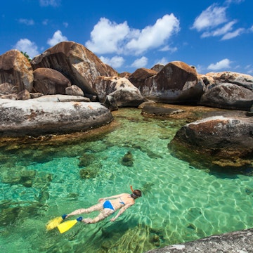 Young woman snorkeling in turquoise tropical water among huge granite boulders at The Baths beach.