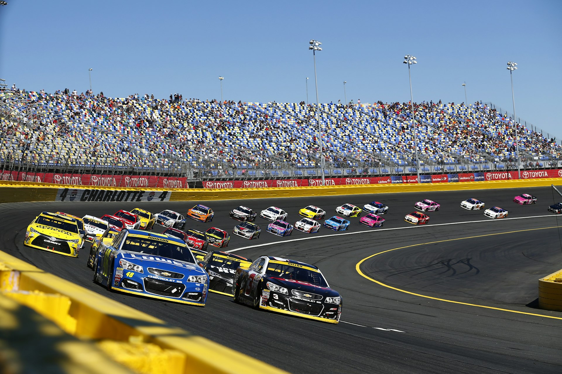 A race track with cars speeding round and the stands filled with spectators. Jimmie Johnson (48) and Austin Dillon (3) lead the field to a restart during the Bank of America 500 at the Charlotte Motor Speedway in Concord.