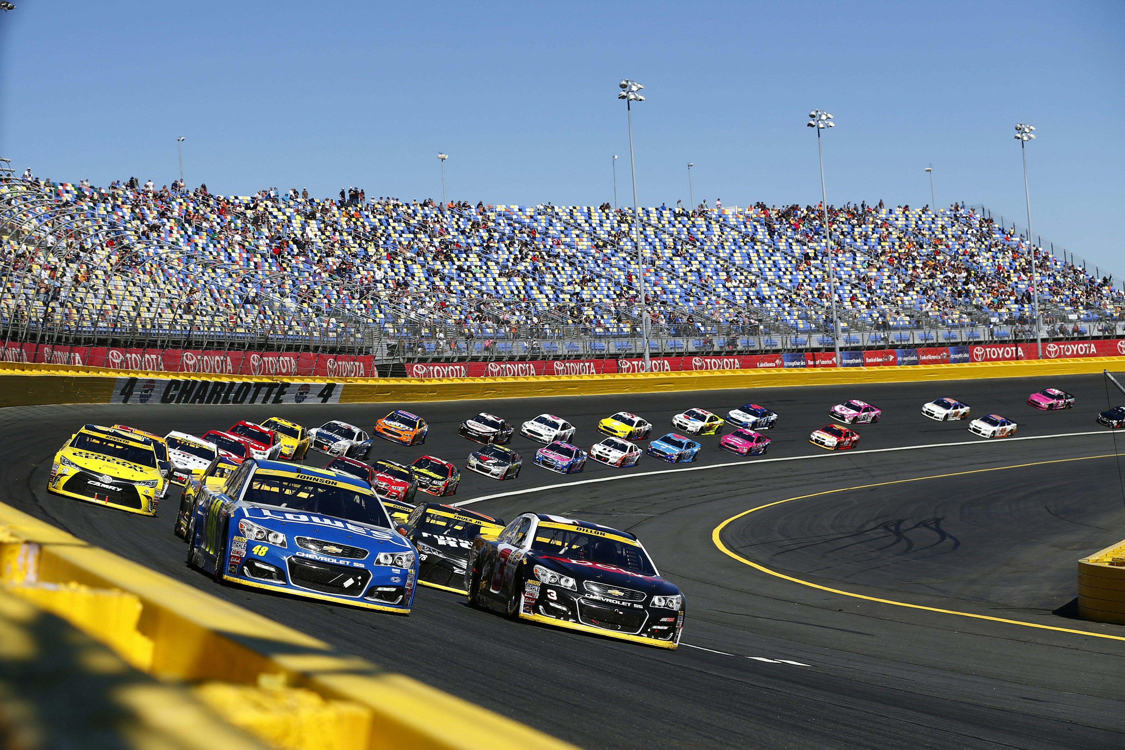 A race track with cars speeding round and the stands filled with spectators. Jimmie Johnson (48) and Austin Dillon (3) lead the field to a restart during the Bank of America 500 at the Charlotte Motor Speedway in Concord.