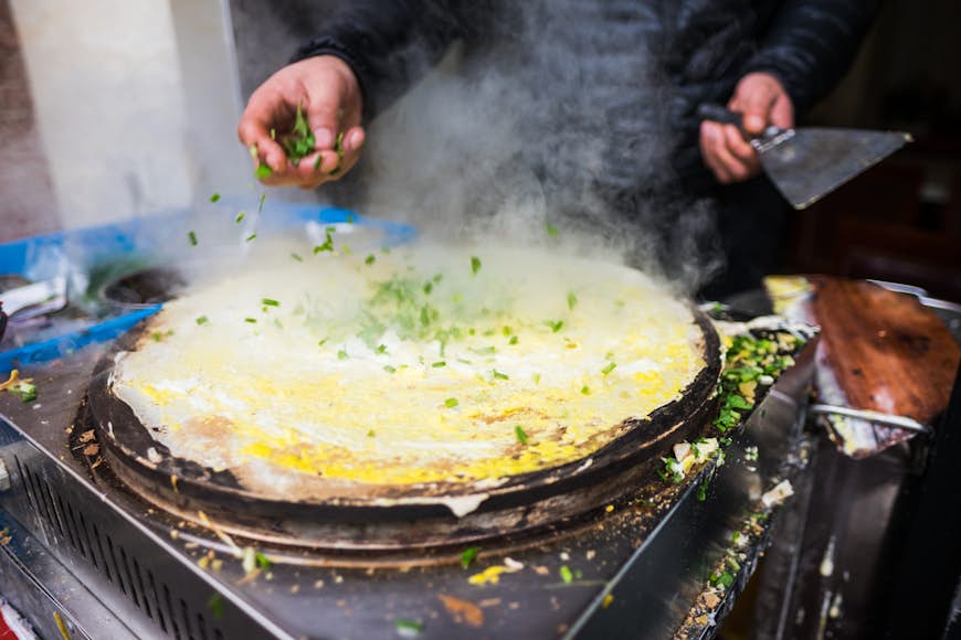 En gatukock sprider salladslök över smeten på en stekpanna för att göra en välsmakande jianbing