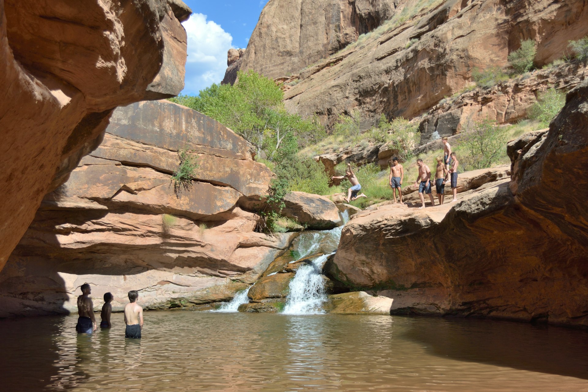 Several swimmers about to jump off a cliff by a waterfall to join other bathers in a swimming hole at Millcreek Canyon