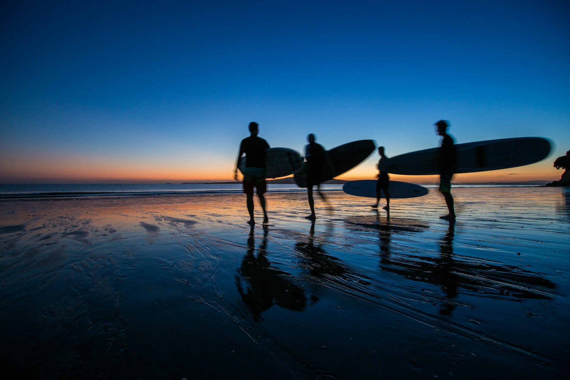 Surfers silhouetted on the beach at dawn with a pink-and-blue sunrise at Takapuna Beach 