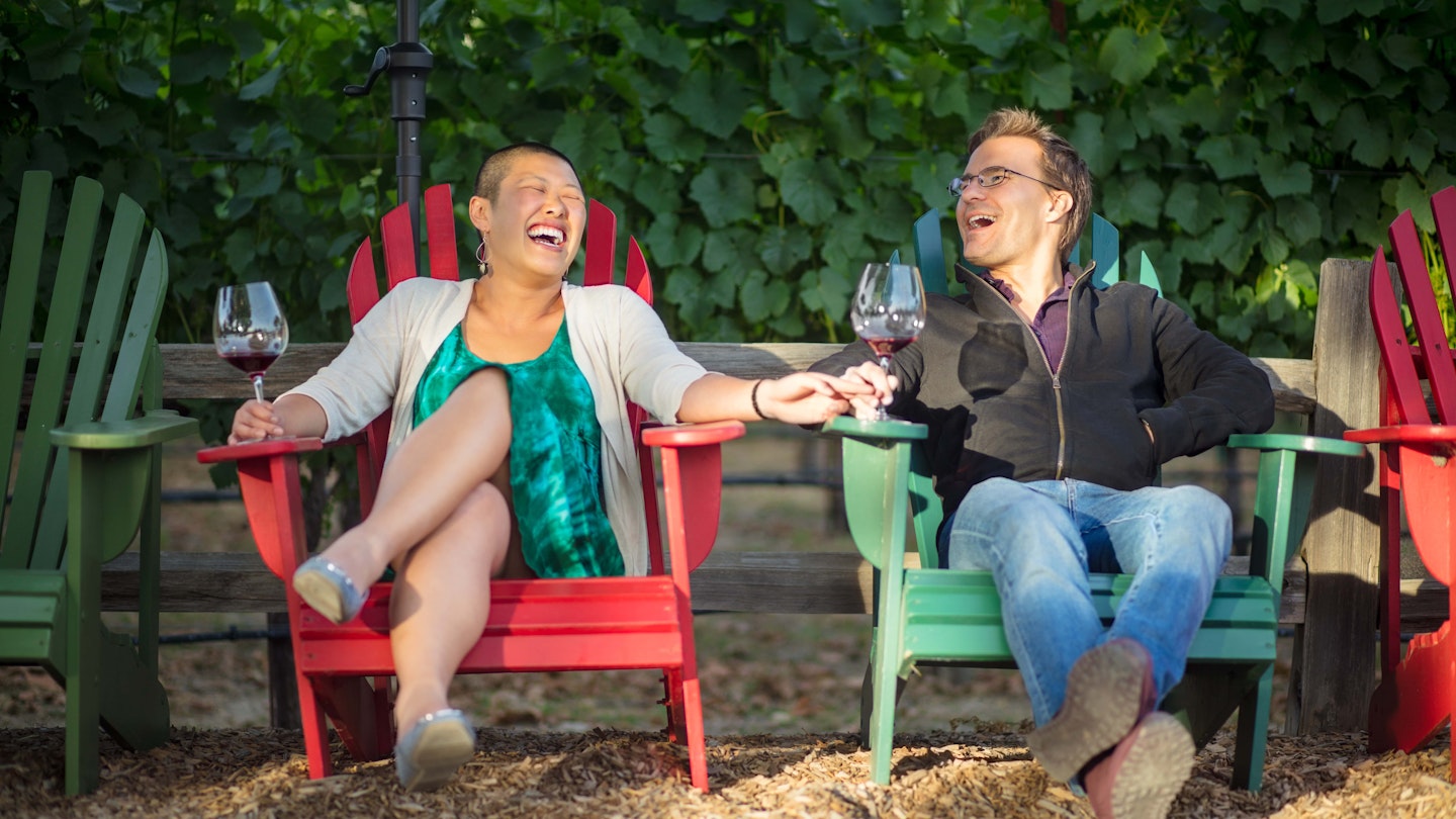 A couple sit in colourful deck chairs in a California vineyard, laughing and holding wine glasses.