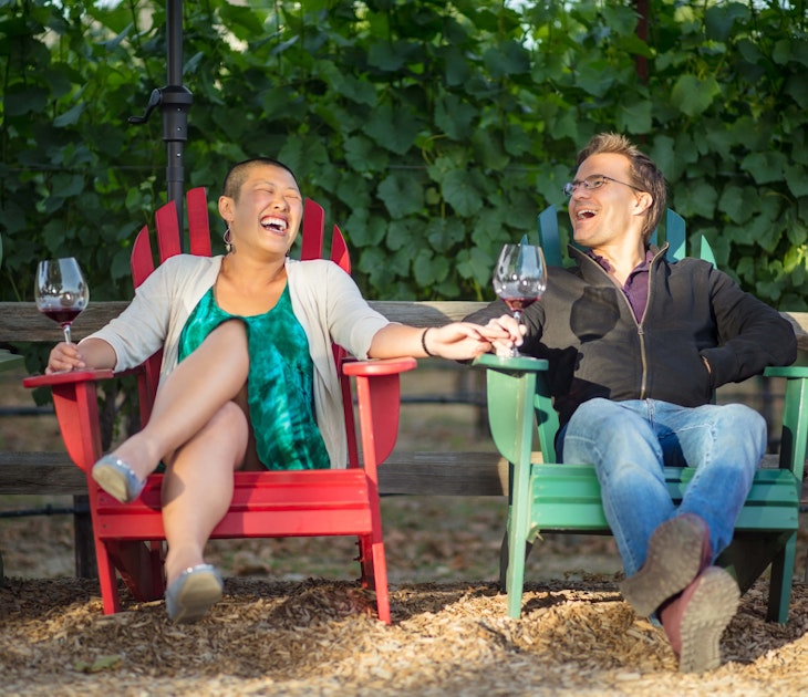 A couple sit in colourful deck chairs in a California vineyard, laughing and holding wine glasses.