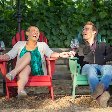 A couple sit in colourful deck chairs in a California vineyard, laughing and holding wine glasses.