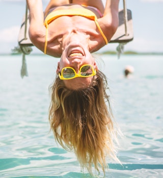 Woman playing on swing over the sea in Mexico, she is wearing yellow sunglasses that match her yellow bathing suit and she is smiling joyously. 