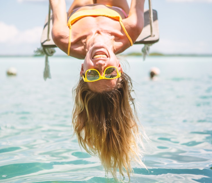 Woman playing on swing over the sea in Mexico, she is wearing yellow sunglasses that match her yellow bathing suit and she is smiling joyously. 