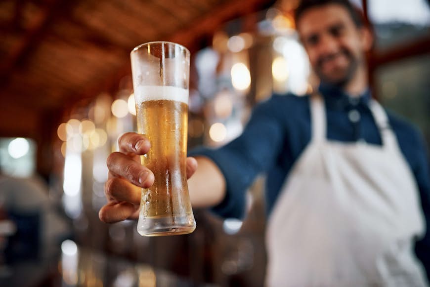 Närbild av en glad ung företagsägare som håller upp ett glas öl som han precis hällt upp, taget i ett bryggeri under dagen