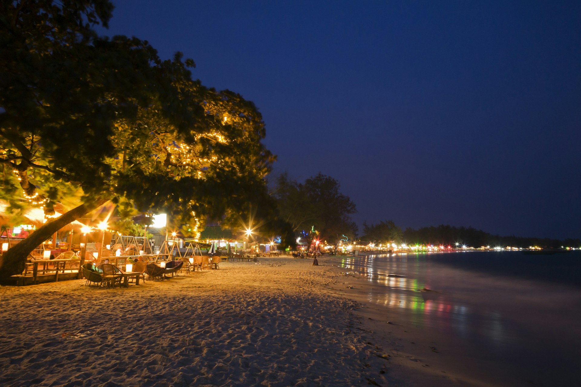 An empty sandy beach at night, lit by the lights from beachside bars.