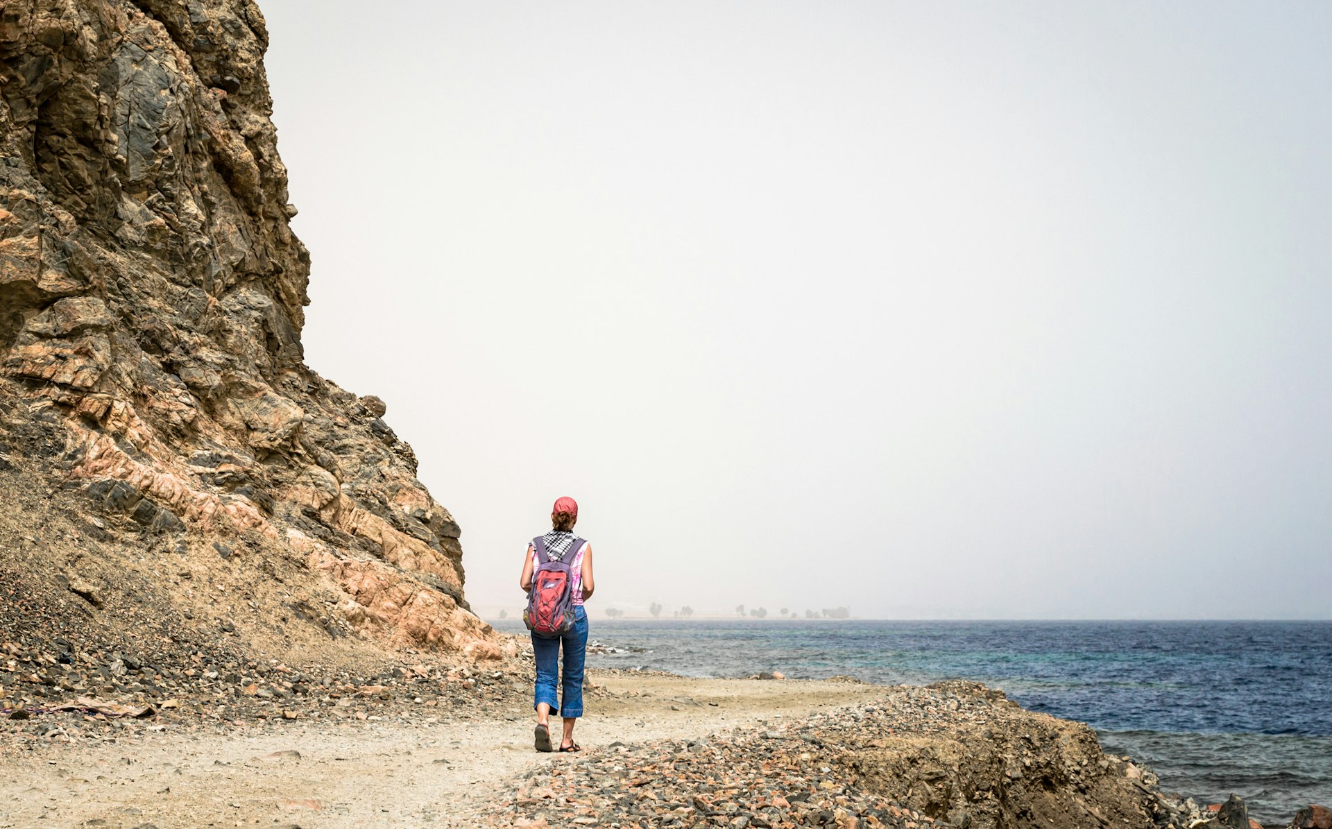 young woman tourist in a red cap with a backpack and sandals hikes along the rocky shore of the Red Sea in Egypt