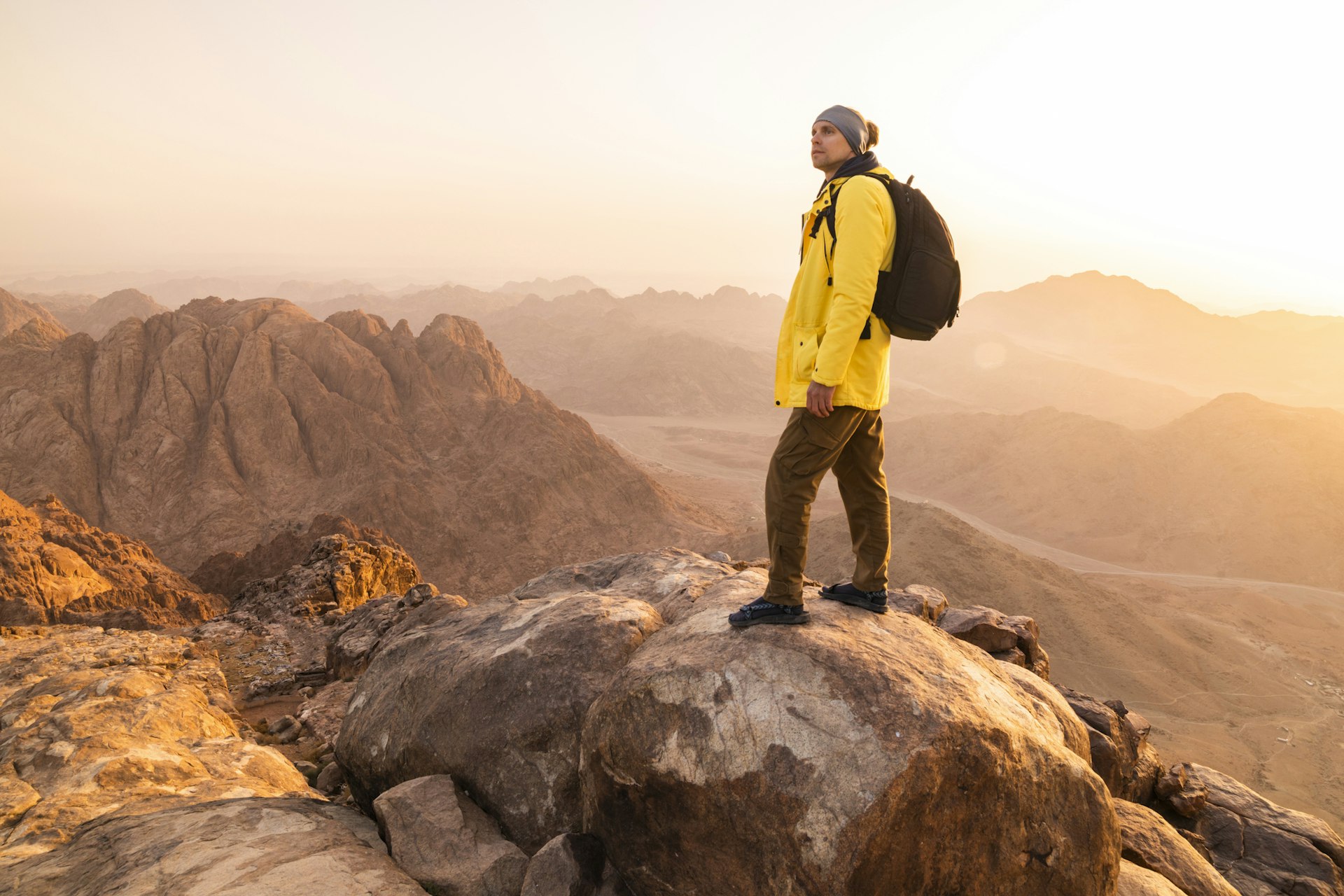 A hiker in a bright yellow coat with a black rucksack stands at the top of a mountain in Sinai, Egypt at sunset 