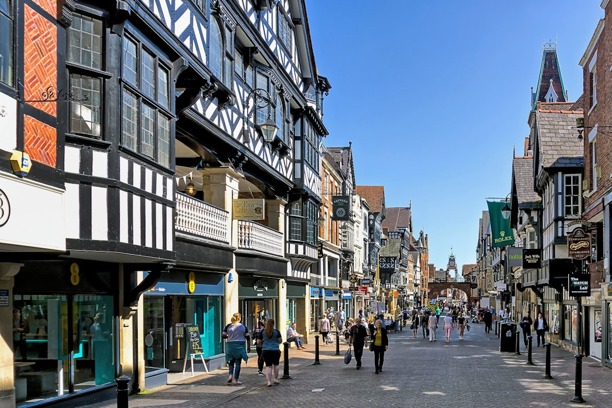 View along the main street in the centre of Chester, Cheshire, UK.  Shops can be seen on either side of the road and people can be seen walking and sitting on benches. The rows of upper level shops are pictured on left. 
