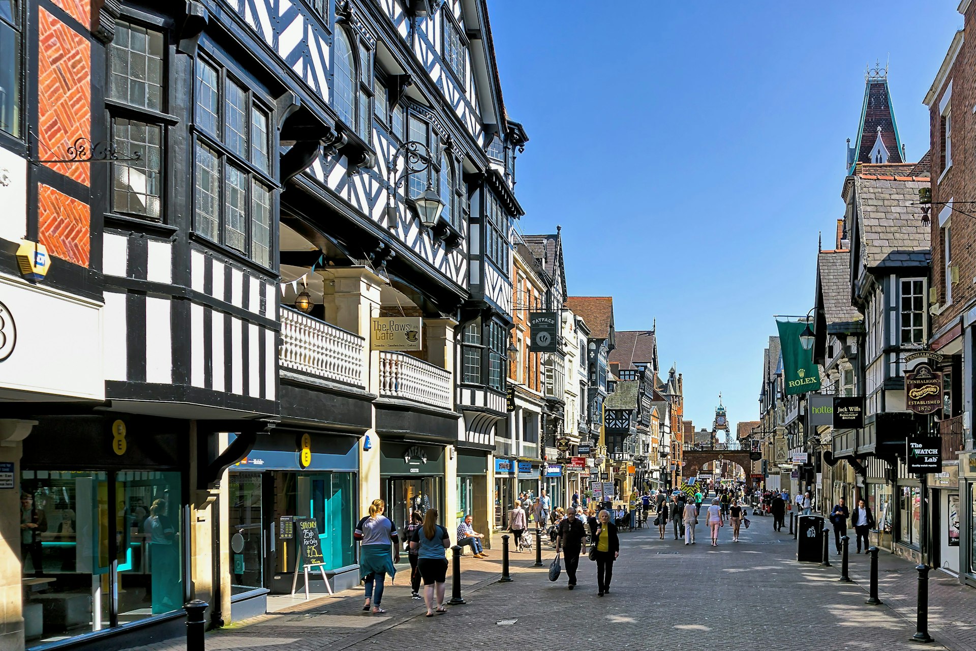 View along the main street in the centre of Chester, Cheshire, UK.  Shops can be seen on either side of the road and people can be seen walking and sitting on benches. The rows of upper level shops are pictured on left. 