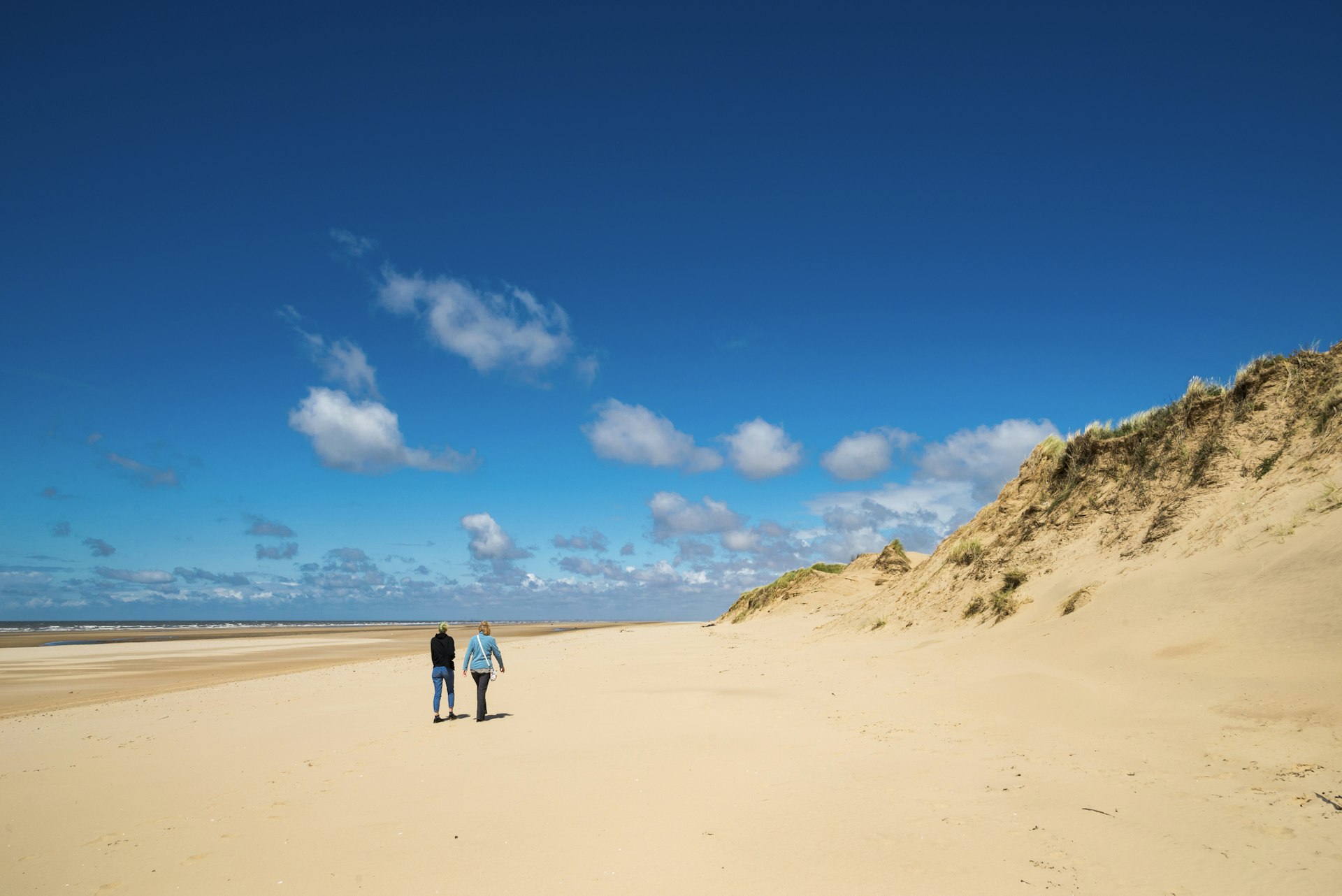 Two people walk along a vast empty sandy beach, with sea to their left and sand dunes to their right