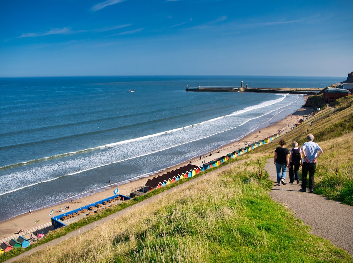 Tourists on a path overlooking colourful beach huts, the coast and harbour piers at Whitby, North Yorkshire, UK - taken on a sunny day at the end of summer