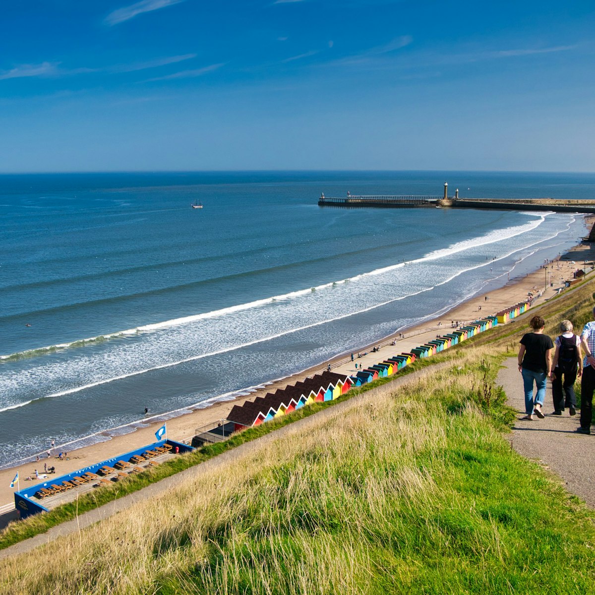 Tourists on a path overlooking colourful beach huts, the coast and harbour piers at Whitby, North Yorkshire, UK - taken on a sunny day at the end of summer