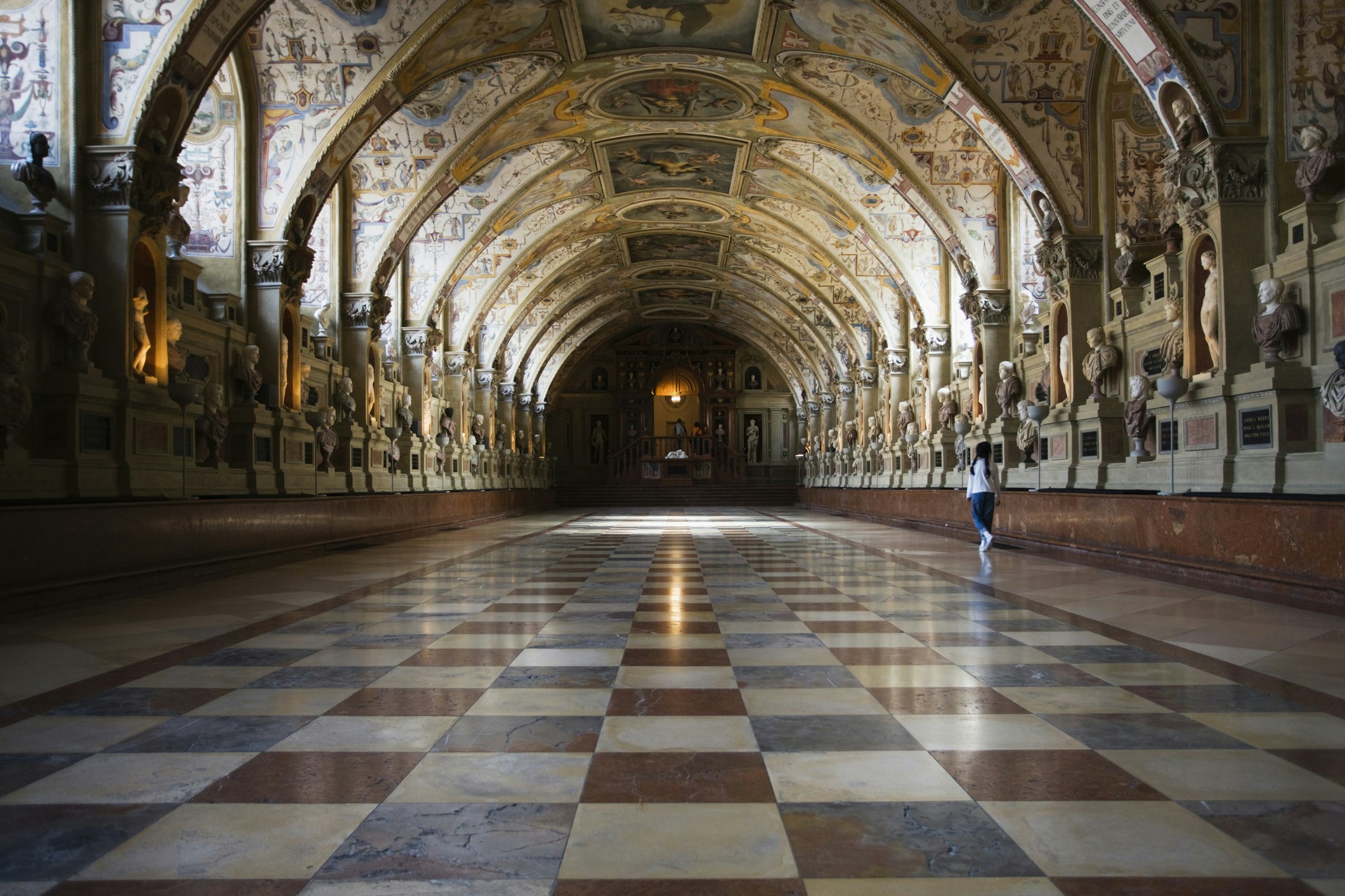 A long wide hallway with a checked tiled floor. The vaulted ceiling is covered in paintings