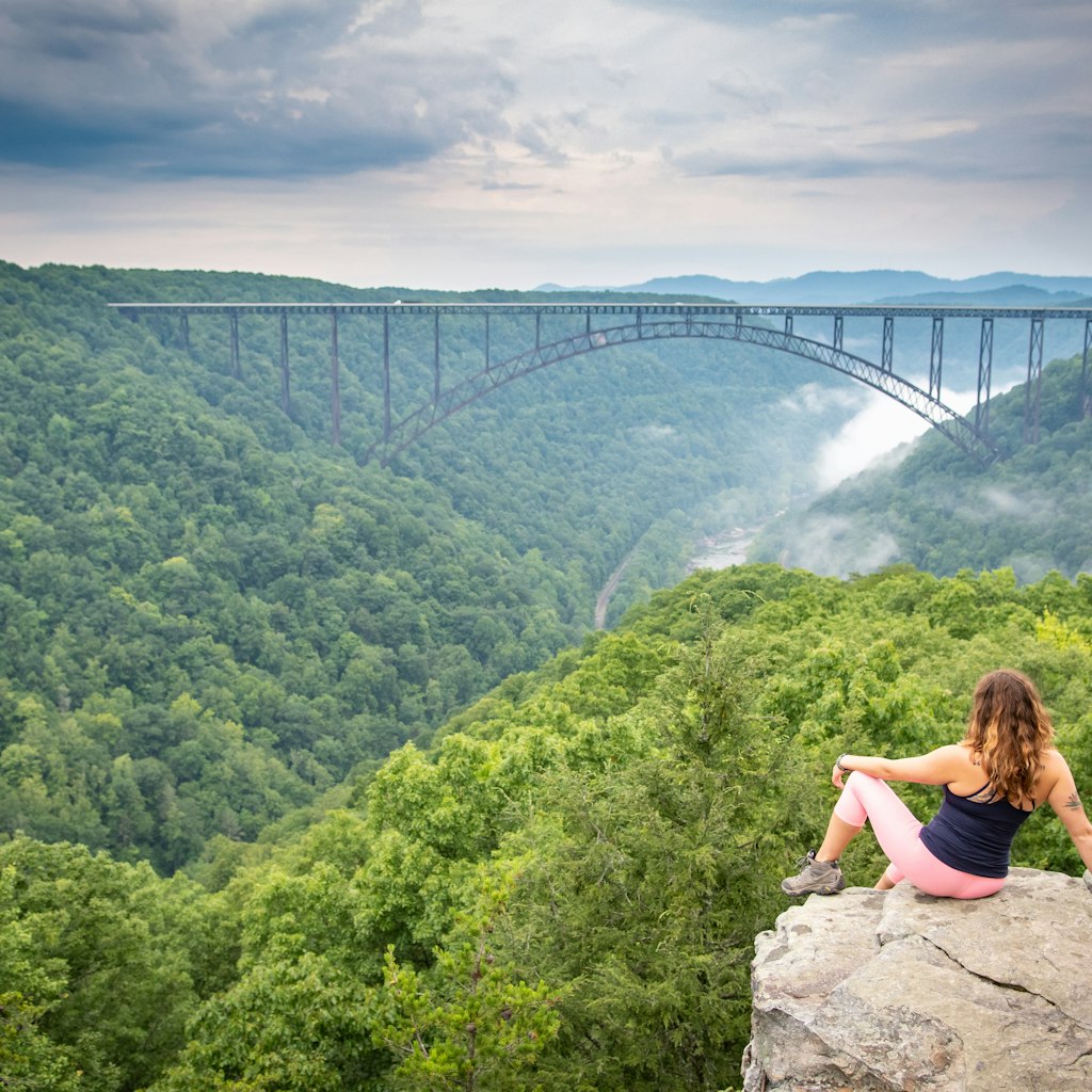 A woman perched on a rocky outcrop looks out over the New River Gorge Bridge in Wild West Virginia.  A moody sky and foggy river below add drama.