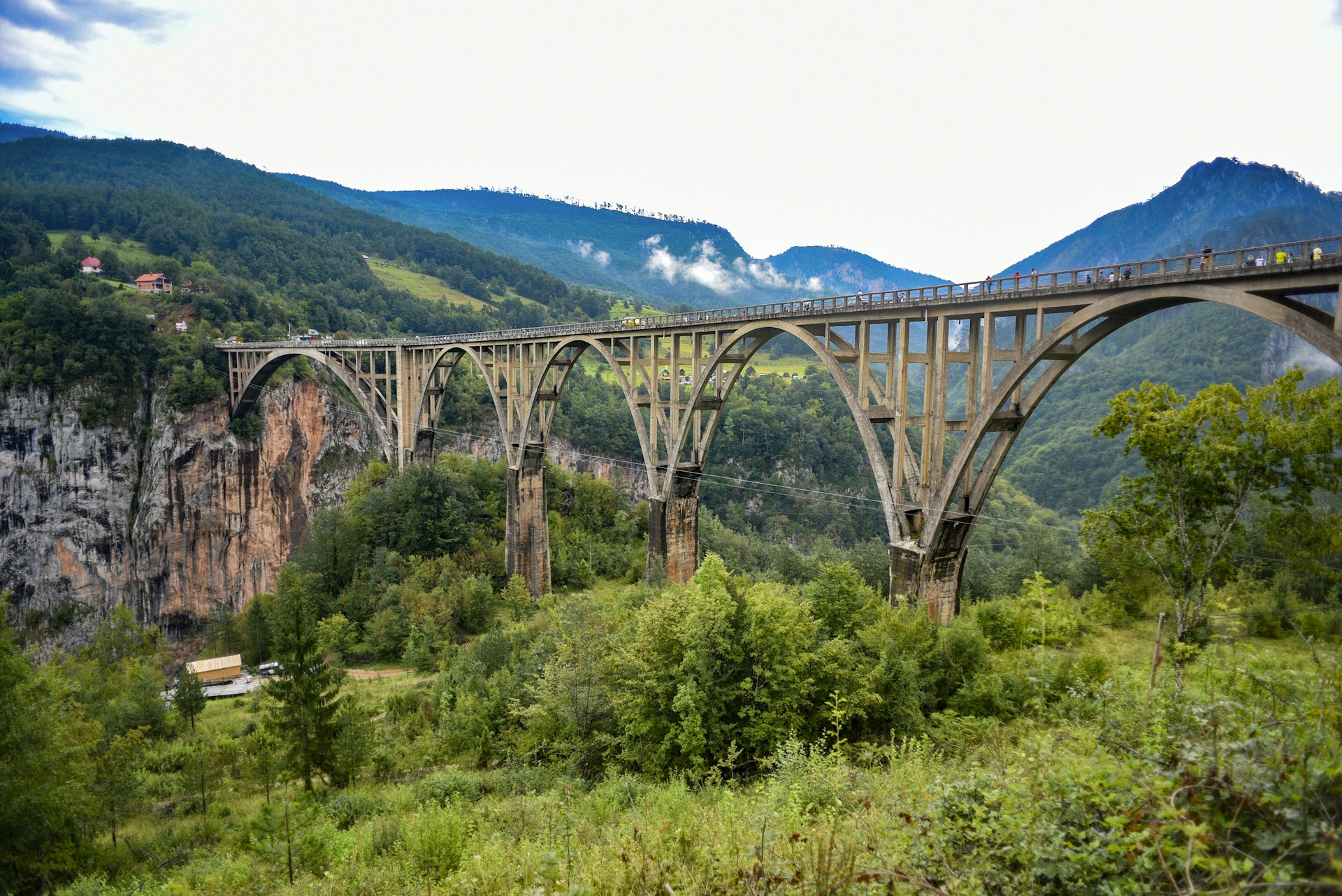 Pessoas caminham por uma ponte com vários arcos que atravessa um desfiladeiro em uma área montanhosa