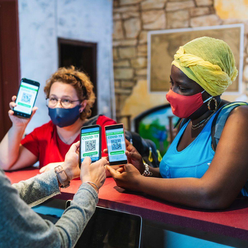 Friends with backpacks and wearing protective face mask arriving to a city hostel, welcomed by female receptionist who checks their digital certificate of Covid-19 vaccination passport