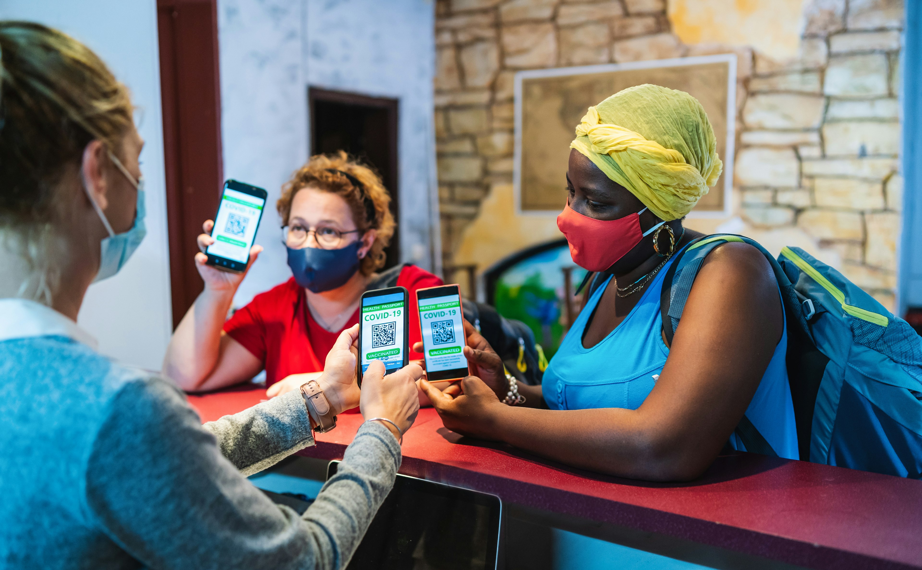 Friends with backpacks and wearing protective face mask arriving to a city hostel, welcomed by female receptionist who checks their digital certificate of Covid-19 vaccination passport