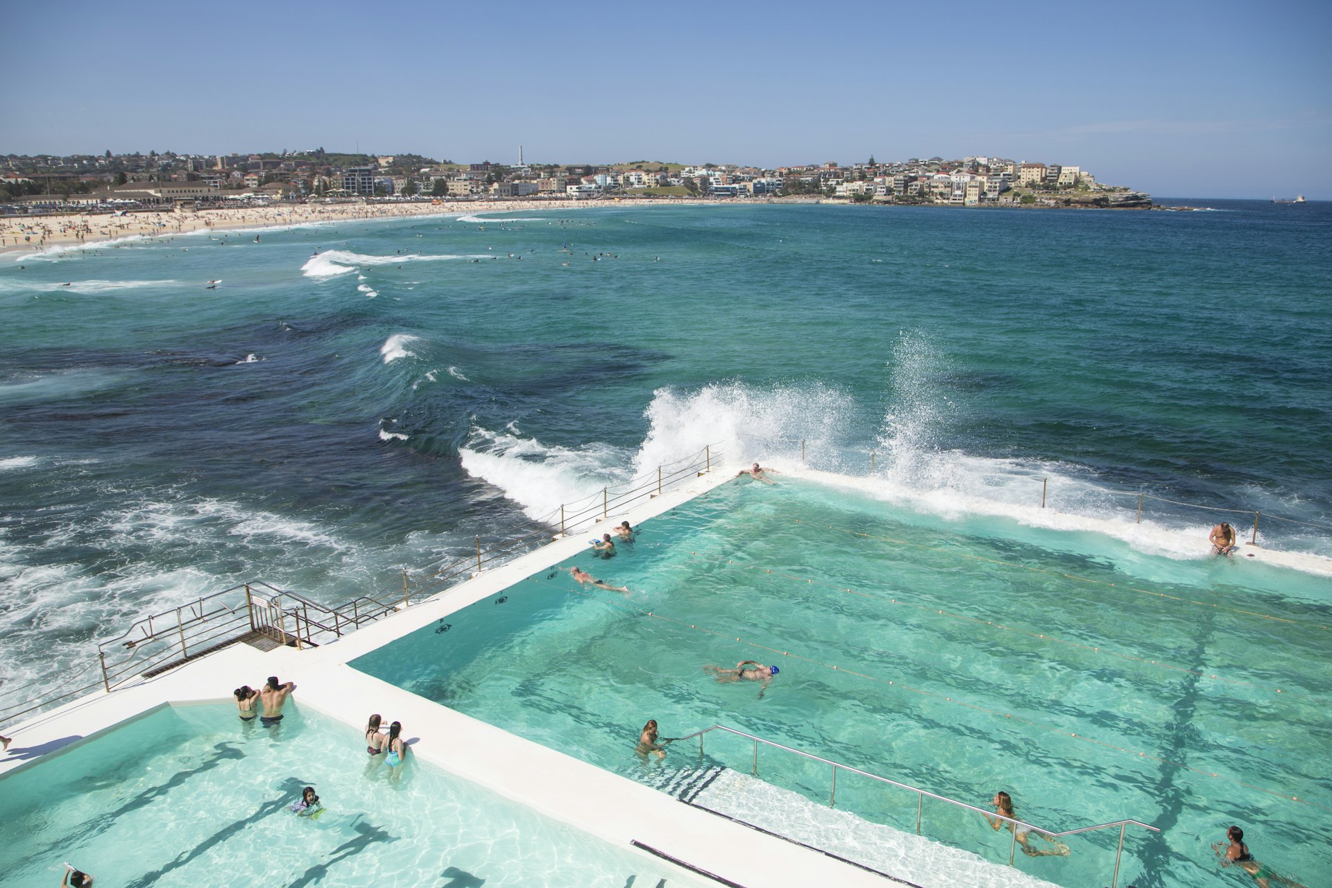 People swimming in the freshwater swimming pools built in to the sea with waves rolling in to Bondi and breaking against the edge of the pool