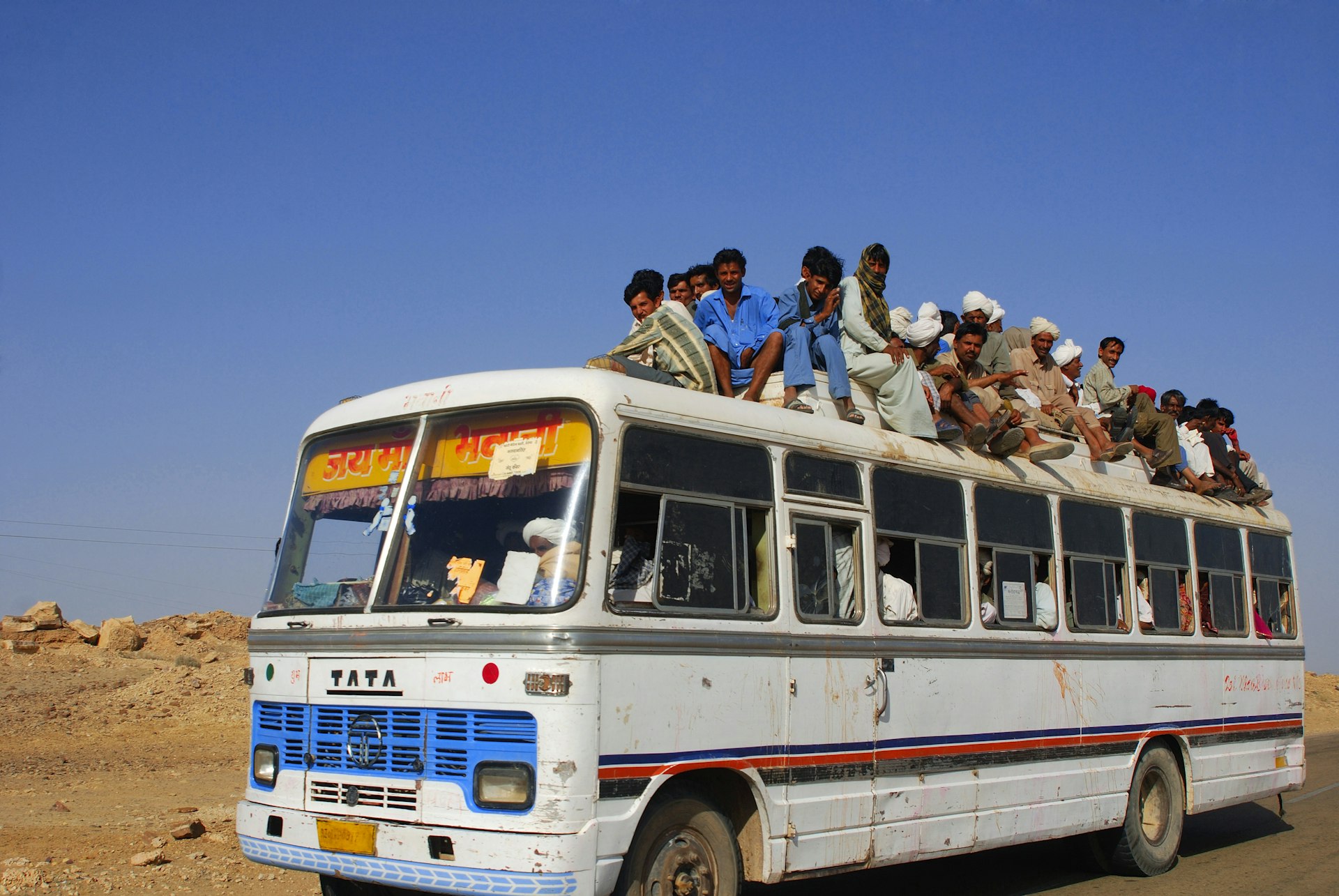 People crowding on the roof of a bus as it drives through an arid landscape