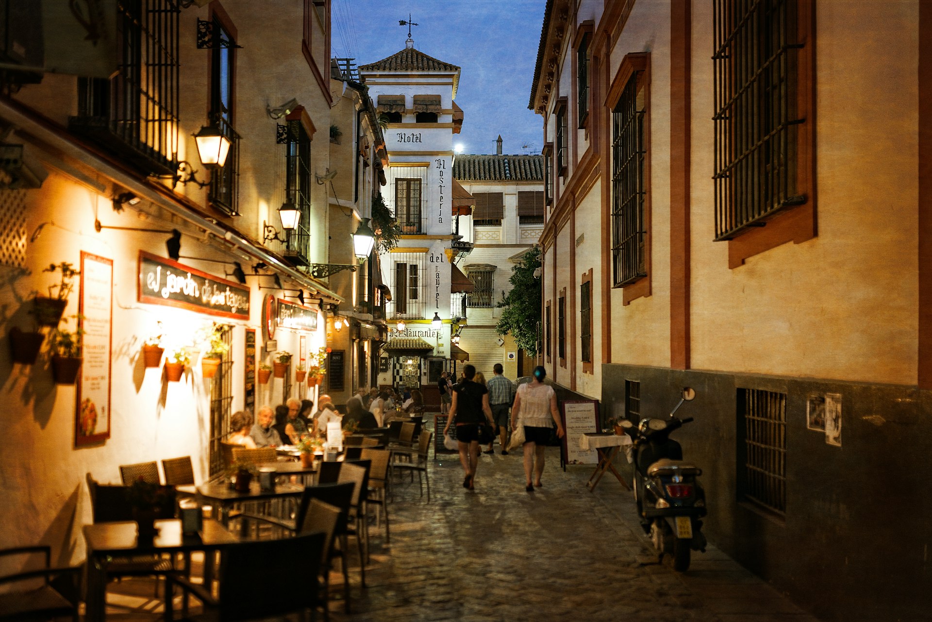 People walk by tables on an outdoor terrace of a restaurant in a narrow side street in Seville, Spain