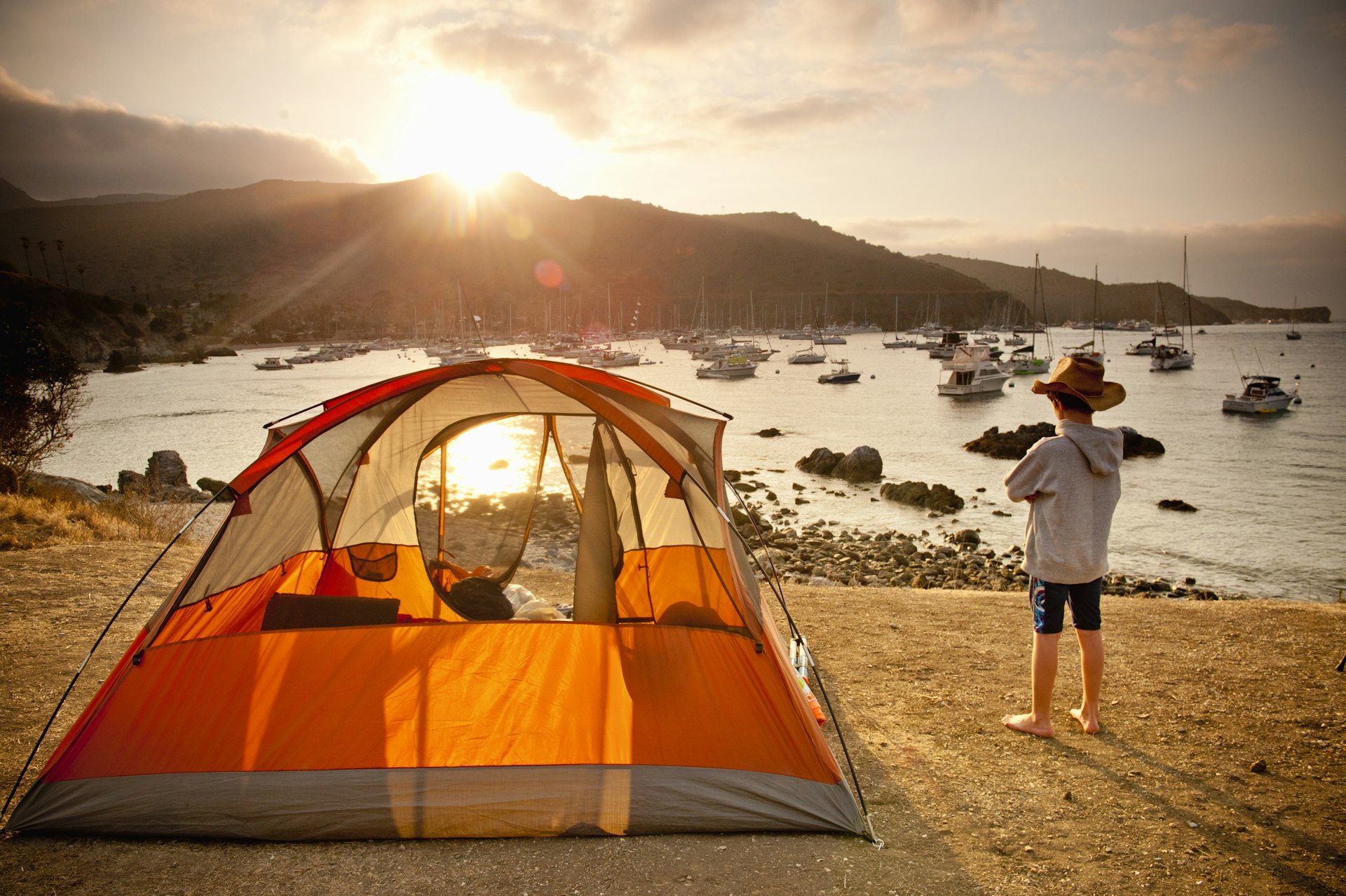 A child stands outside a tent overlooking a harbor as the sun rises