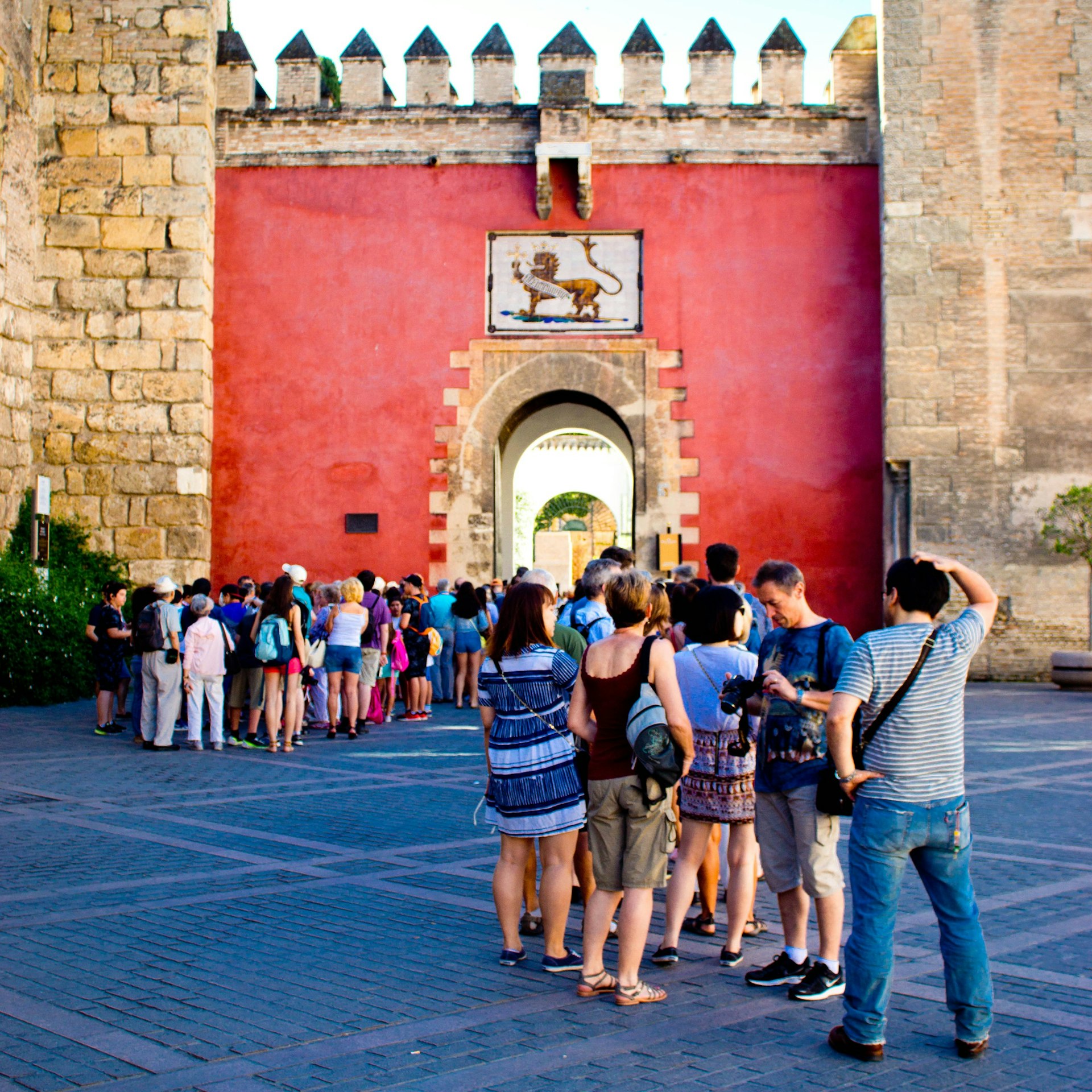 The exterior wall of the Real Alcázar castle in Spain with a crest of a lion on the wall below the castellated top of the wall. In the foreground are tourists queuing for entry