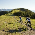 Two young men ride their mountain bikes down a singletrack trail in the Rocky Mountains of Canada. They are both riding enduro-style mountain bikes and are wearing hydration backpacks.