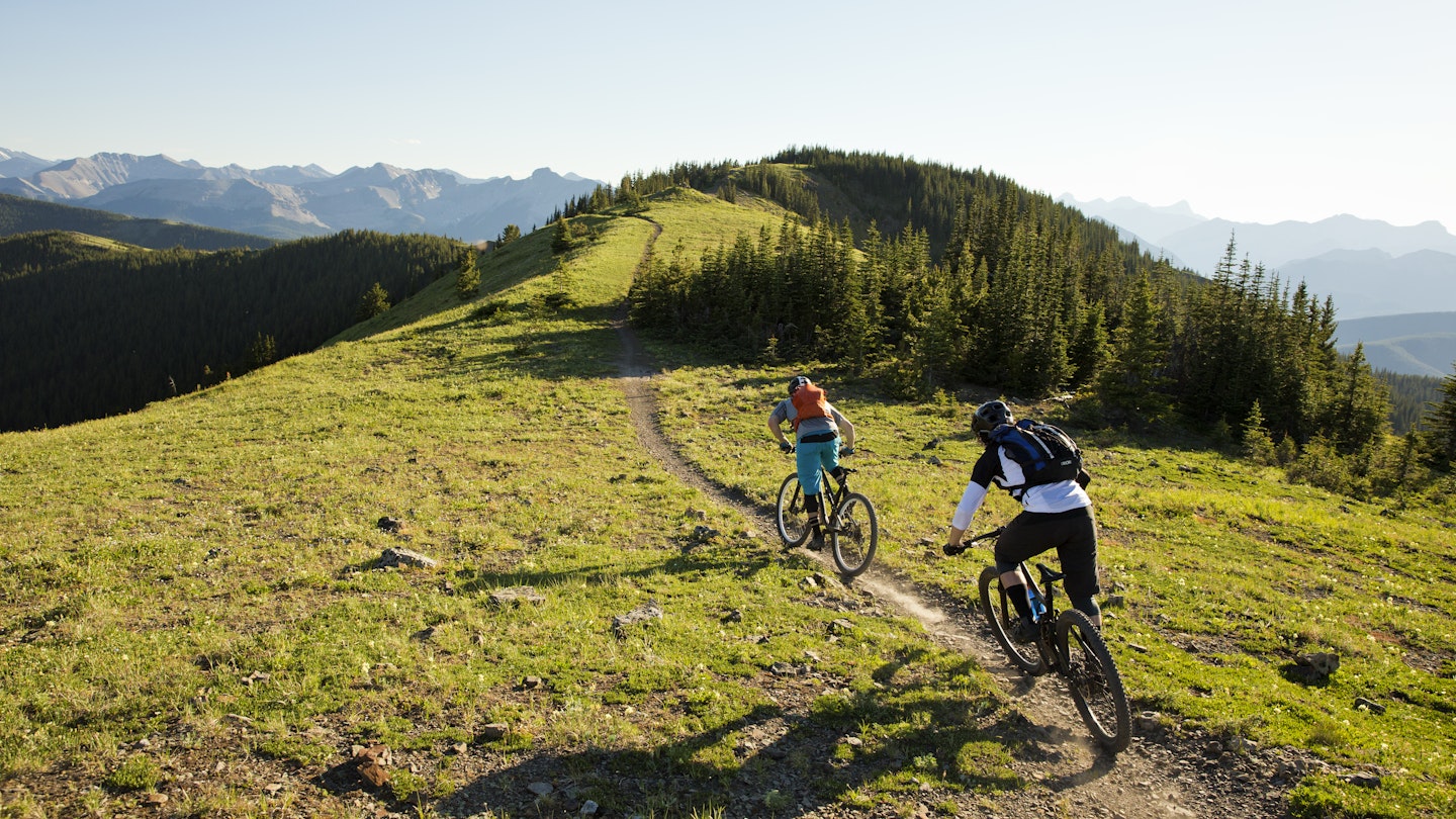 Two young men ride their mountain bikes down a singletrack trail in the Rocky Mountains of Canada. They are both riding enduro-style mountain bikes and are wearing hydration backpacks.