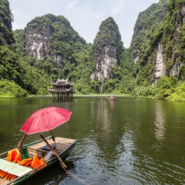 Empty sampan with a red umbrella surrounded by steep limestone cliffs and with a small temple in the background.