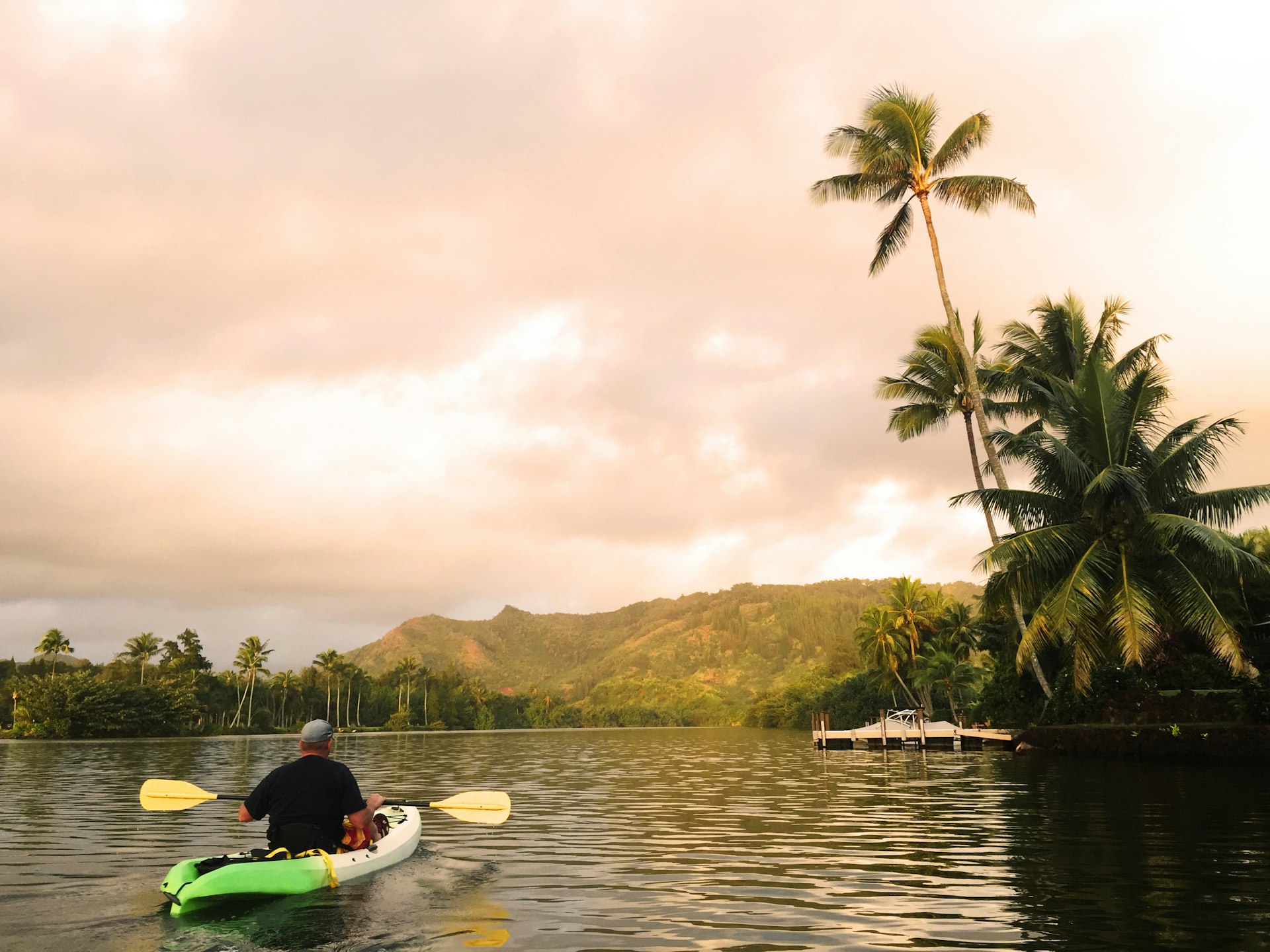 A man kayaks on Wailua River as early morning light turns the sky a soft pink with palms on the right
